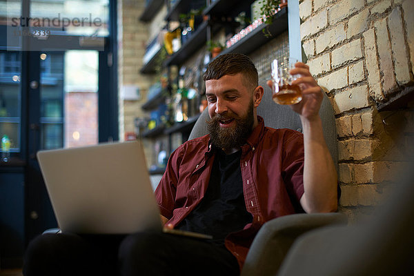 Junger Mann mit Becher des Geistes schaut auf den Laptop im Wirtshaus