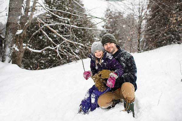 Porträt eines mittelgroßen Mannes  der im Schnee kauert und seine Tochter umarmt