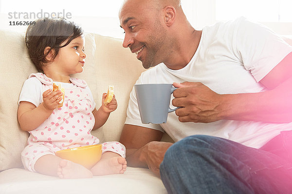 Kleines Mädchen und Vater sitzen auf dem Sofa und essen einen Snack
