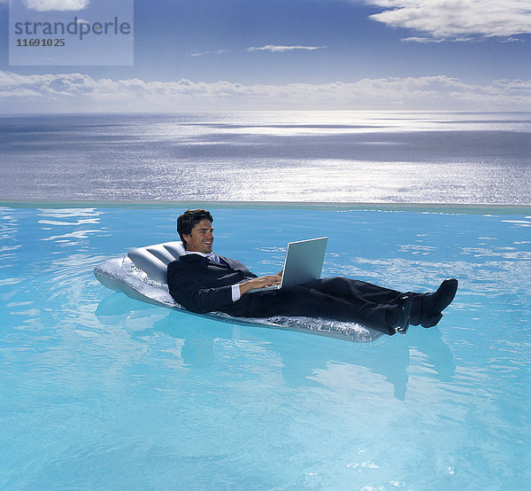 Ein Mann im Anzug auf einer Luftmatratze im Schwimmbad  ein Laptop auf seinem Schoß.