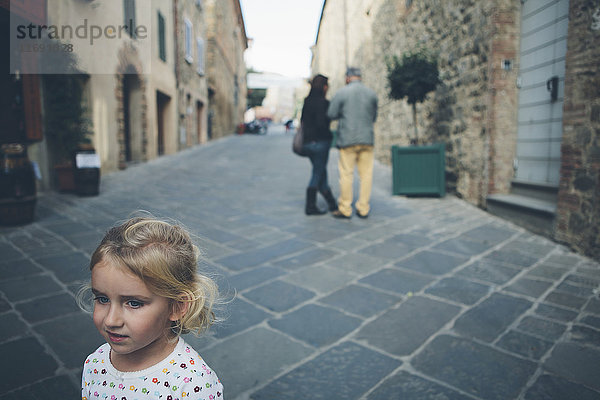 Ein dreijähriges Mädchen allein auf einer Straße in einer Stadt  mit zwei Erwachsenen im Hintergrund  die sich den Rücken zugewandt haben.