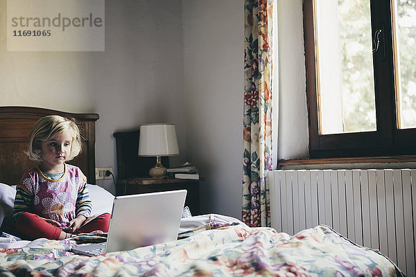 Ein dreijähriges Mädchen sitzt auf einem Bett in einem Hotelzimmer und schaut aufmerksam auf einen Laptop-Computerbildschirm.