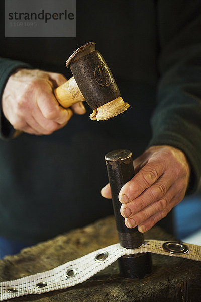 Nahaufnahme eines Mannes in der Werkstatt eines Segelmachers  der Ösen in einen Segeltuchstreifen hämmert.