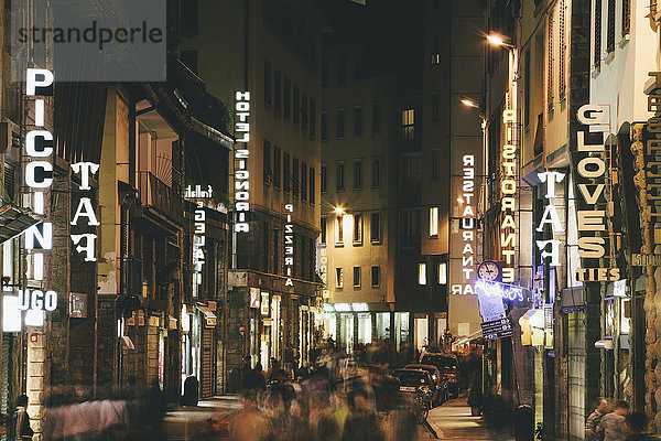 Eine Straße in Florenz wird nachts mit Neonschildern beleuchtet  und historische Gebäude erleuchten. Lange Belichtung. Laufende Menschen