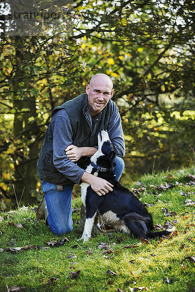 Schafzüchter  der auf einer Wiese kniend seinen schwarz-weißen Schäferhund tätschelt.