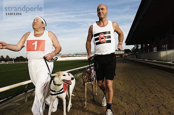 Zwei Männer in Sportkleidung laufen auf einer Rennstrecke mit zwei Windhunden an der Leine.