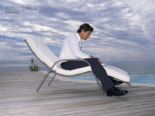 Ein Mann in weißem Hemd und schwarzer Hose in einem Liegestuhl mit einem Laptop auf seinem Schoß.