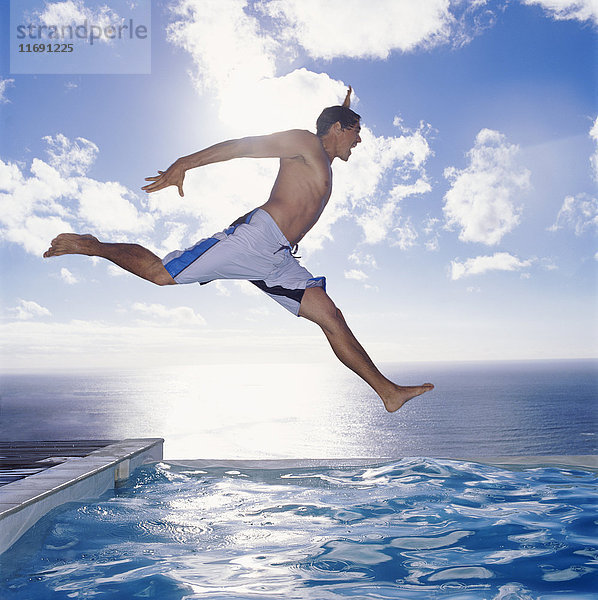 Mann in Badehose  der in ein Schwimmbad am Meer  am Himmel und an den Wolken springt.