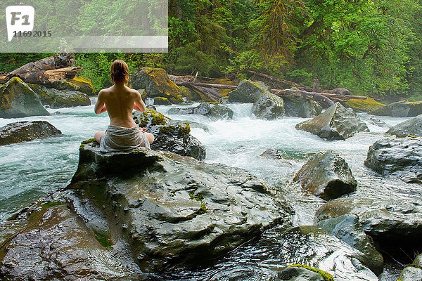 Teilweise bekleidete Frau meditiert auf Felsen am Wasser