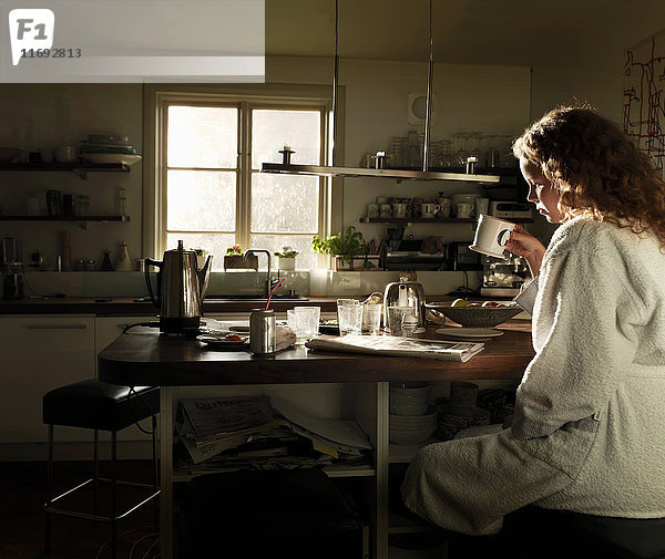 Junge Frau liest Zeitung am Küchentisch beim Frühstück