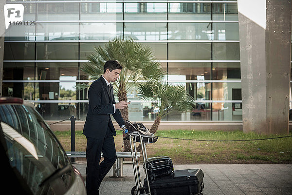 Junger Mann benutzt Mobiltelefon mit Gepäckwagen am Flughafen