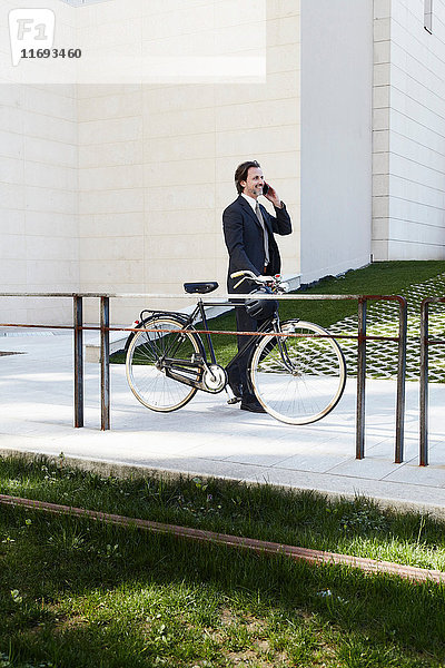 Mittlerer erwachsener Geschäftsmann  der ein Fahrrad hält und ein Mobiltelefon benutzt
