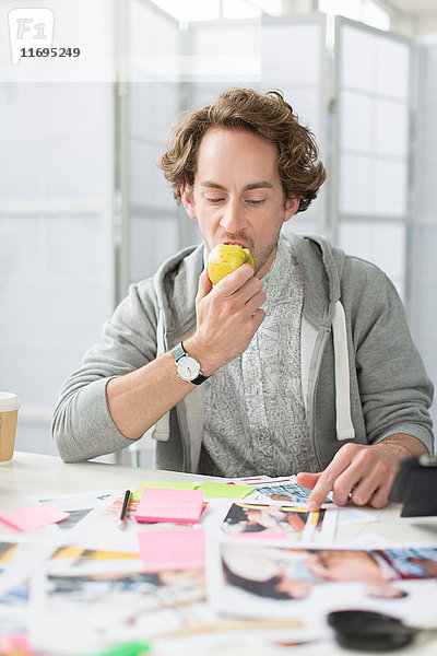 Junger Mann isst Apfel am Schreibtisch im Kreativbüro