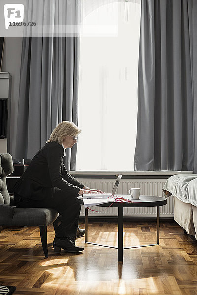 Seitenansicht der reifen Geschäftsfrau mit Laptop am Fenster im Hotelzimmer