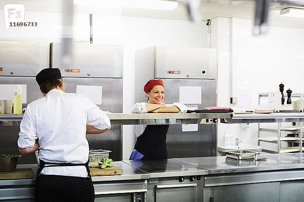 Lächelnde Köchin  die sich auf den Tresen stützt  mit Schülerin  die in der Kochschule arbeitet
