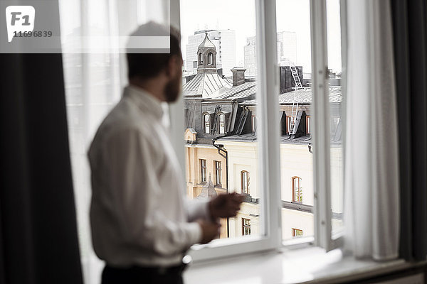 Seitenansicht eines Geschäftsmannes  der durch ein Fenster vom Hotelzimmer aus auf die Gebäude schaut.
