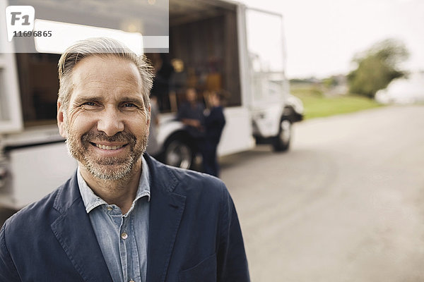 Portrait eines glücklichen Geschäftsmannes auf der Straße mit Kollegen und tragbarem Bürowagen im Hintergrund