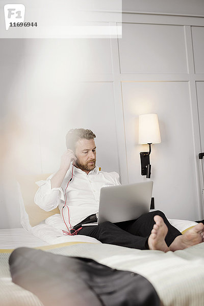Ein reifer Geschäftsmann  der im Hotelzimmer auf dem Bett sitzt und den Kopfhörer am Laptop einstellt.