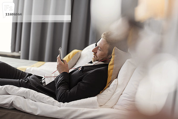 Seitenansicht eines Geschäftsmannes  der Musik hört  während er im Hotelzimmer auf dem Bett liegt.