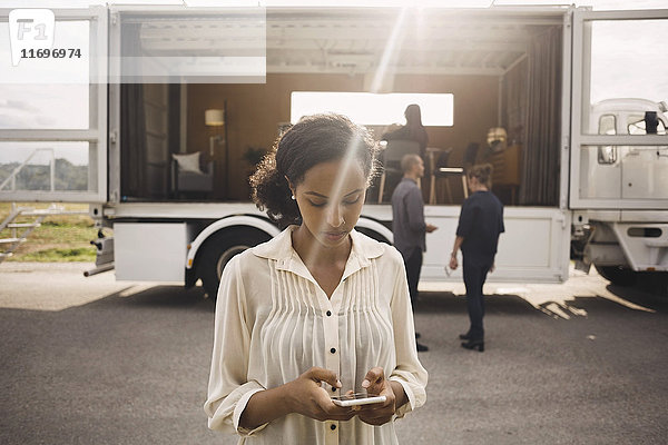 Geschäftsfrau mit Smartphone mit Kollegen und tragbarem Bürowagen im Hintergrund