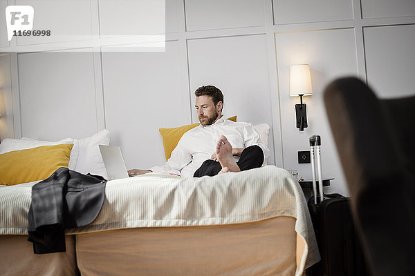 Geschäftsmann auf dem Bett sitzend mit Laptop an der Wand im Hotelzimmer