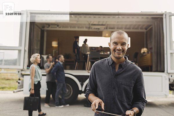 Porträt eines lächelnden Geschäftsmannes  der im Hintergrund ein digitales Tablett mit Kollegen und einem tragbaren Bürowagen auf der Straße hält.