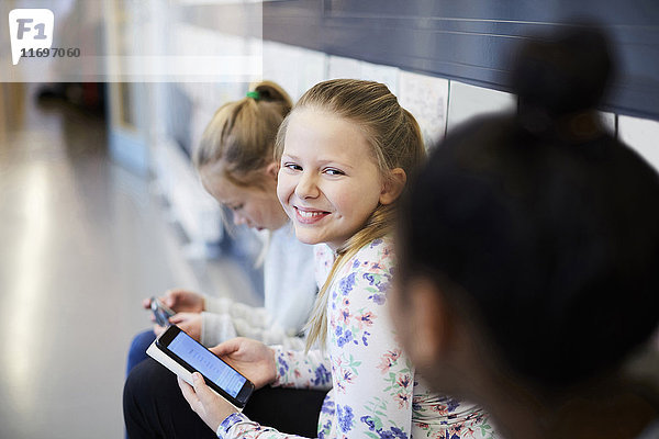 Lächelndes Mädchen hält Smartphone  während es einen Freund im Schulflur ansieht.