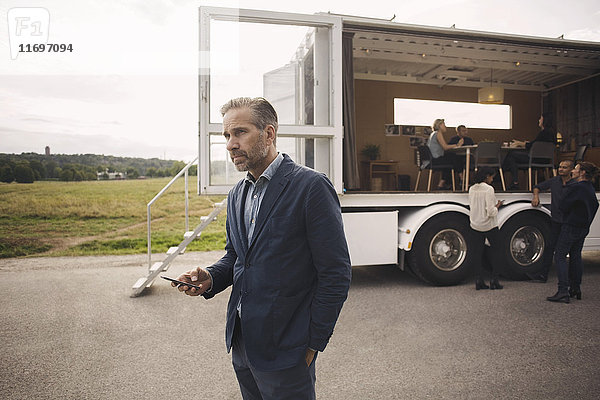Geschäftsmann hält Handy mit Kollegen und tragbarem Bürowagen im Hintergrund