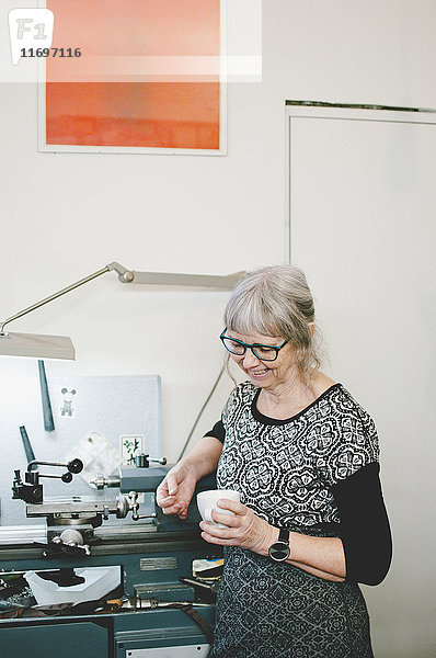 Seniorin lächelt  während sie eine Kaffeetasse in der Schmuckwerkstatt hält.