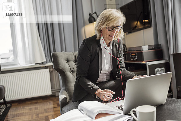 Reife Geschäftsfrau  die Kopfhörer hört  während sie den Laptop im Hotelzimmer benutzt.