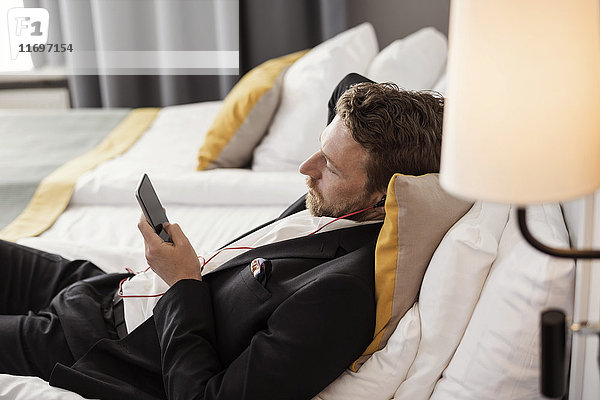 Hochwinkelansicht eines Geschäftsmannes  der mit dem Handy Musik hört  während er im Hotelzimmer liegt.
