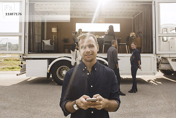 Porträt eines Geschäftsmannes  der sein Handy mit Kollegen und einem tragbaren Bürowagen im Hintergrund hält.