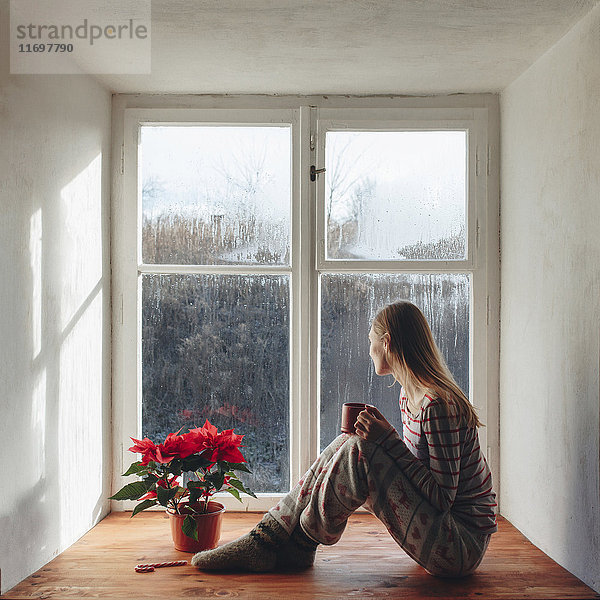Kaukasische Frau sitzt auf der Fensterbank und trinkt Kaffee im Schlafanzug