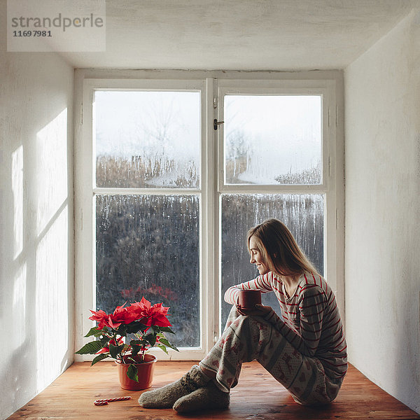 Kaukasische Frau sitzt auf der Fensterbank und trinkt Kaffee im Schlafanzug