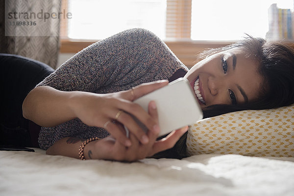 Gemischtrassige Frau liegt auf dem Bett und schreibt eine SMS auf ihrem Handy
