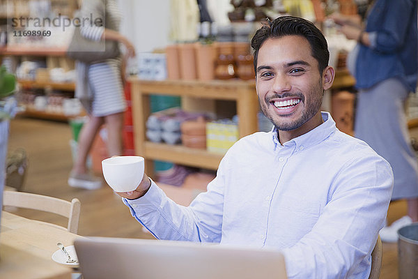 Porträt eines begeisterten jungen Mannes  der in einem Café am Laptop Kaffee trinkt