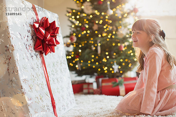 Mädchen lächelt in Erwartung eines großen Weihnachtsgeschenks am Weihnachtsbaum