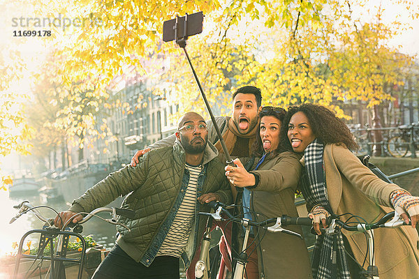 Verspielte junge Freunde mit Fahrrädern  die ein Gesicht mit Selfie-Stick am Herbstkanal machen  Amsterdam