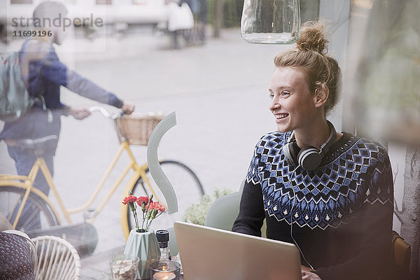 Lächelnde junge Frau mit Kopfhörern  die einen Laptop im Fenster eines städtischen Cafés benutzt