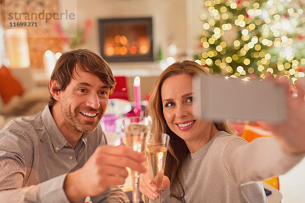 Lächelndes Paar stößt mit Sektflöten an und macht ein Selfie am Weihnachtstisch