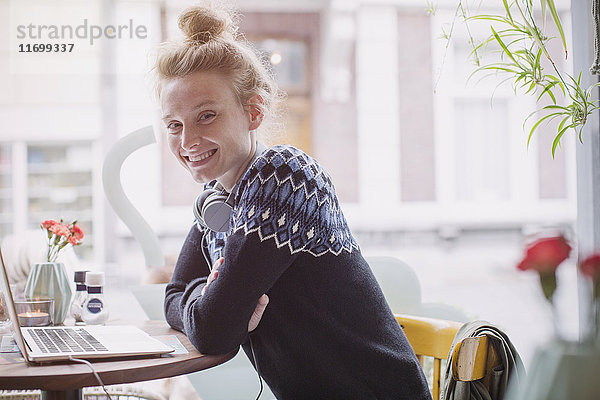 Porträt einer lächelnden jungen Frau mit Kopfhörern am Laptop im Caféfenster