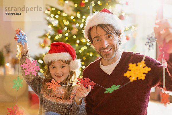 Porträt eines lächelnden Vaters und einer lächelnden Tochter mit Weihnachtsmannmützen  die eine Kette von Papierschneeflocken halten