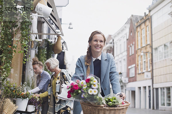 Lächelnde Frau auf dem Fahrrad mit Blumen im Korb auf einer Stadtstraße