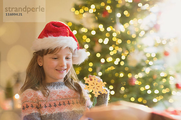 Mädchen mit Weihnachtsmannmütze hält Schneeflockenornament vor einem Weihnachtsbaum