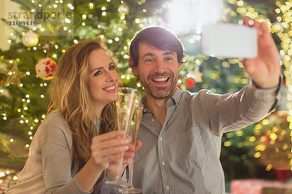 Lächelndes Paar stößt mit Sektflöten an und macht ein Selfie vor dem Weihnachtsbaum
