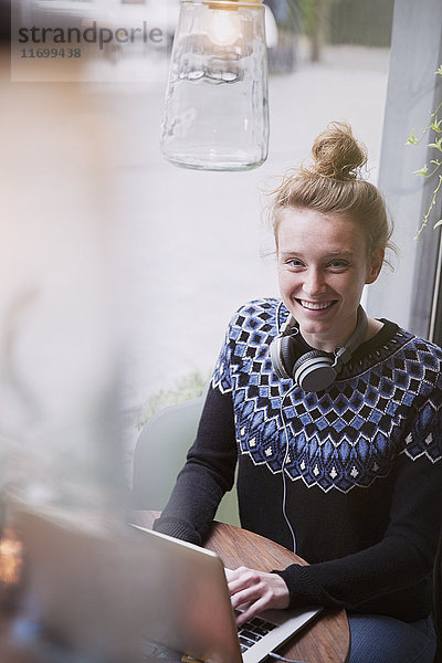 Porträt einer lächelnden jungen Frau  die in einem Caféfenster einen Laptop benutzt