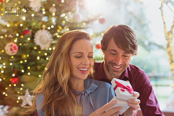 Ehemann beobachtet Frau beim Öffnen von Weihnachtsgeschenken vor dem Weihnachtsbaum