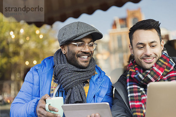 Lächelnde junge Männer Freunde  die Kaffee trinken und Laptop und digitales Tablett im Straßencafé benutzen.