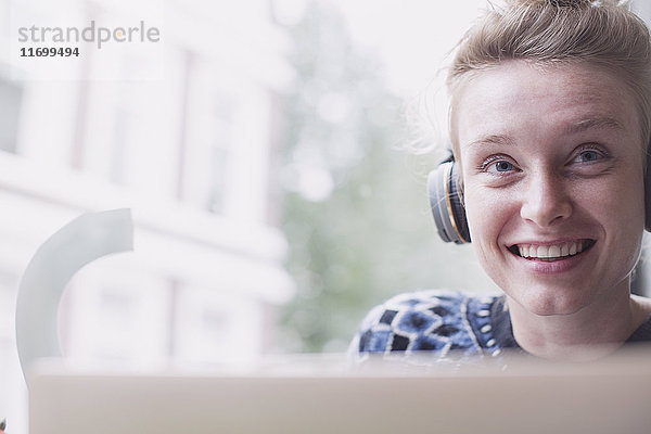 Porträt einer lächelnden jungen Frau mit Kopfhörern am Laptop in einem Caféfenster
