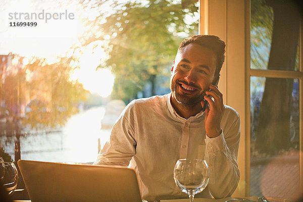 Lächelnder junger Mann beim Telefonieren und Laptopbenutzung im sonnigen Café
